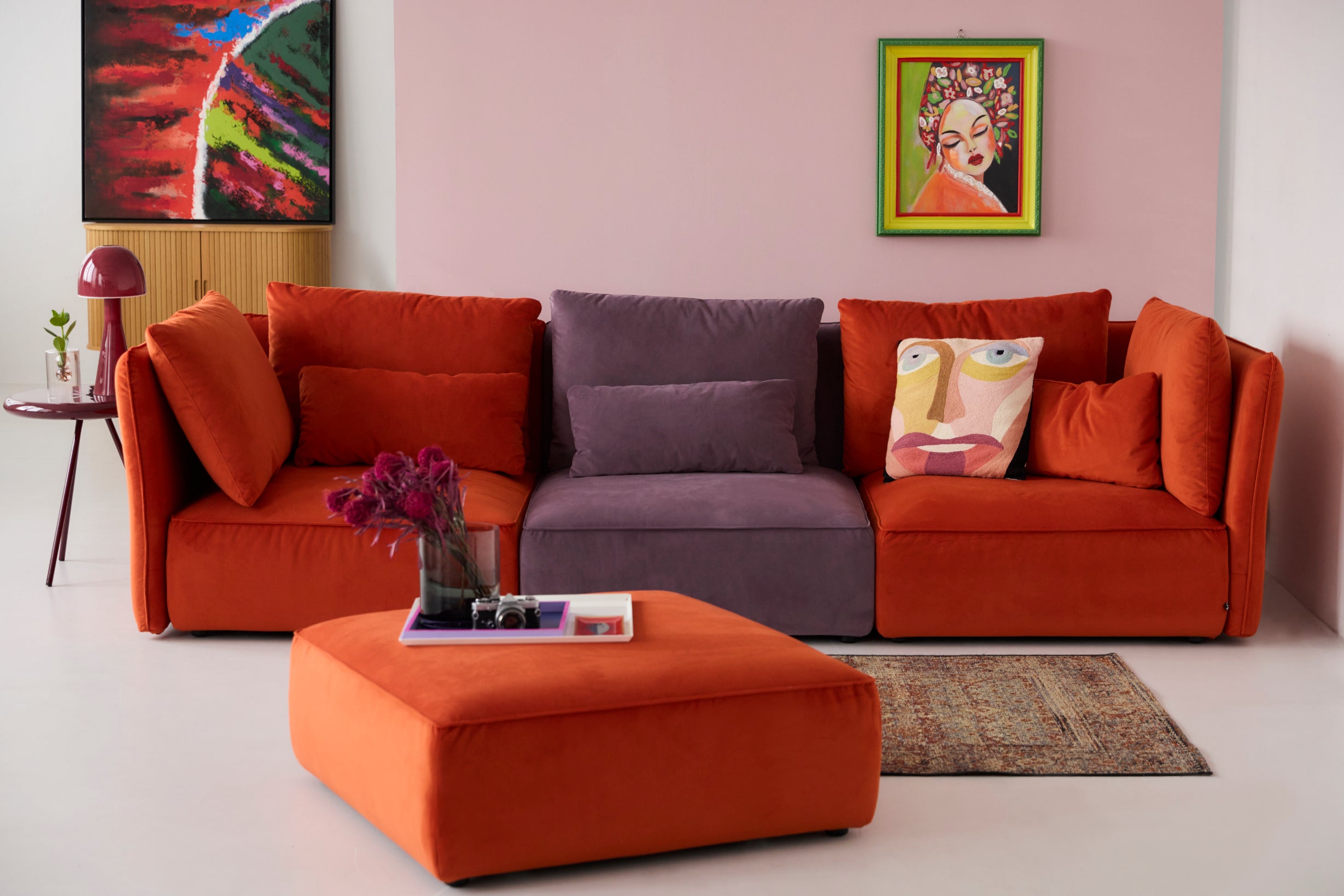 Modulares 3er Sofa NICA von Yvi Living, bestehend aus drei Modulen in kräftigem Orange und sanftem Violett. Die Elemente sind als durchgängige Sofalinie arrangiert und mit passenden Samtkissen dekoriert. Im Vordergrund steht ein passender Samt-Ottoman in Orange, während Wandkunst und Accessoires den Raum akzentuieren. Der Fokus liegt klar auf dem NICA 3er Sofa im Samt und seiner flexiblen Modulbauweise.