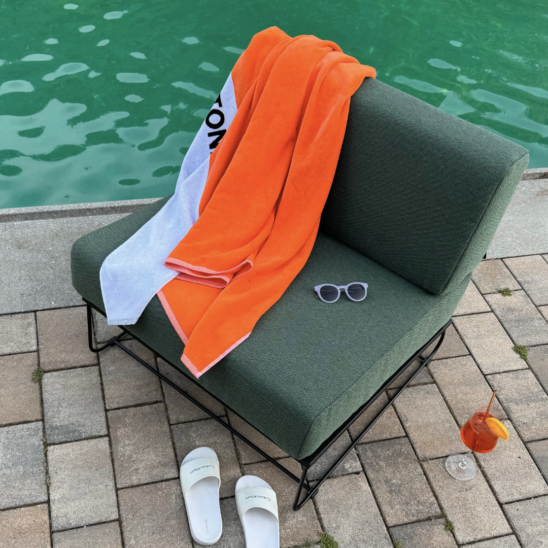Outdoorsessel AKSEL mit dem Stoff Ocean neben einem Pool. Auf dem Outdoorsessel befindet sich ein weiß-oranges Handtuch und eine blaue Sonnenbrille. Vor dem Outdoorsessel befinden sich Schuhe und ein roter Cocktail.