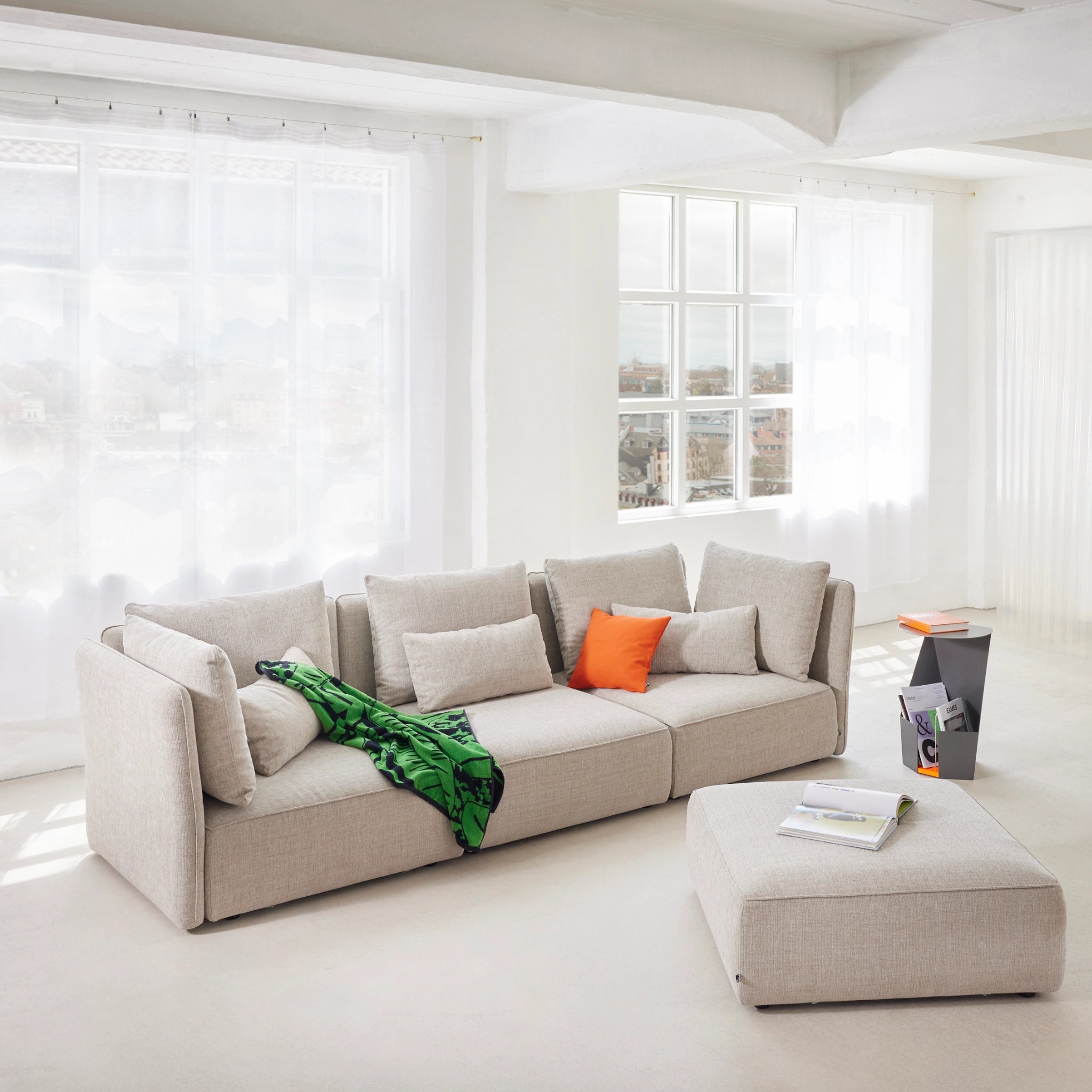 Modernes 3-Sitzer Sofa „NICA“ von Yvi Living in hellem Stoffbezug, inszeniert in einem lichtdurchfluteten Loft mit Pouf, farbigen Kissen und grüner Decke – stilvoller Komfort für ein elegantes, urbanes Wohnambiente.