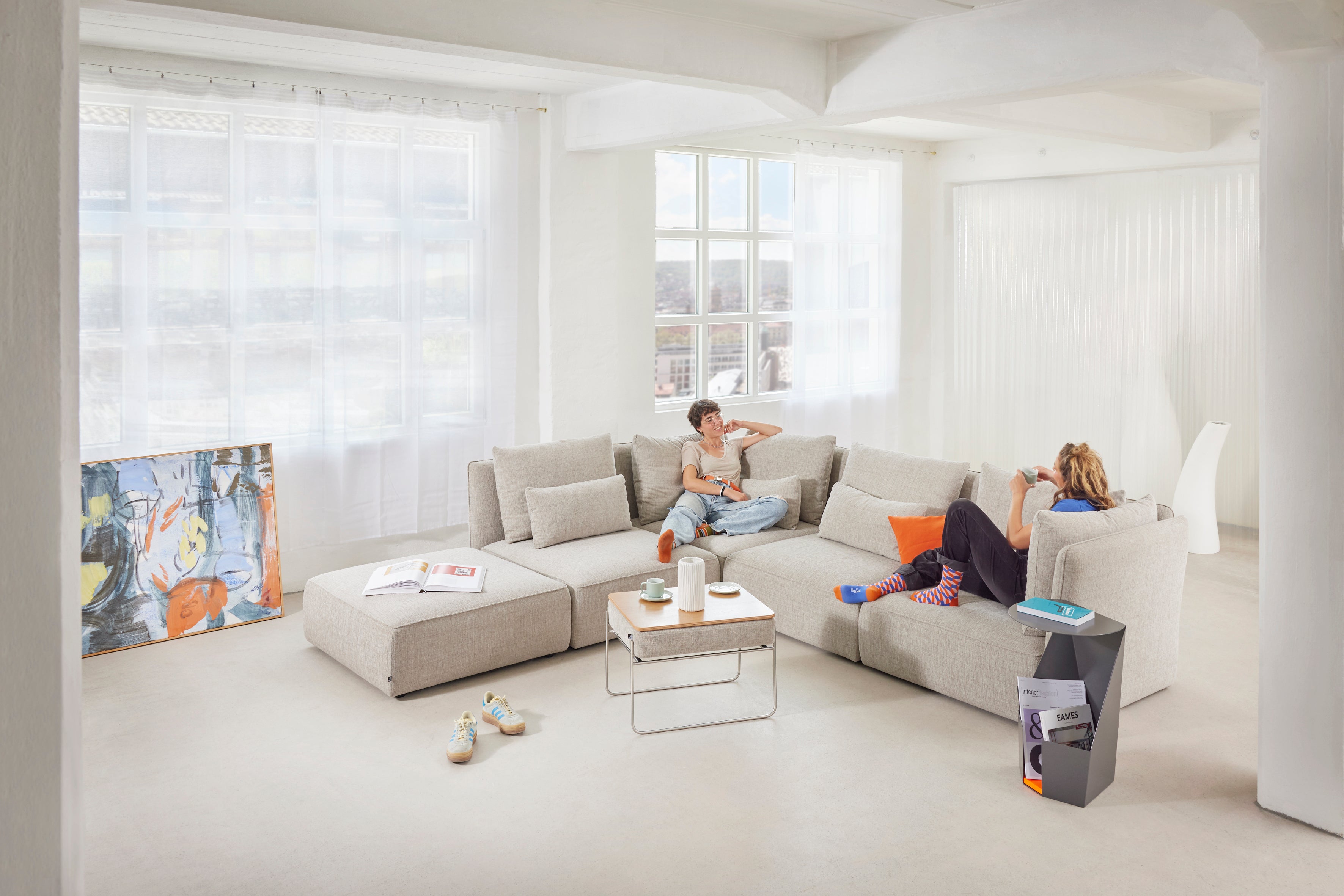Großzügiges modulares NICA Sofa von Yvi Living in hellem Stoff, kombiniert mit Hocker und Beistelltisch – gemütliche Szene mit zwei Personen in einem lichtdurchfluteten Loft. Perfekte Verbindung aus Design, Komfort und entspannter Wohnatmosphäre.