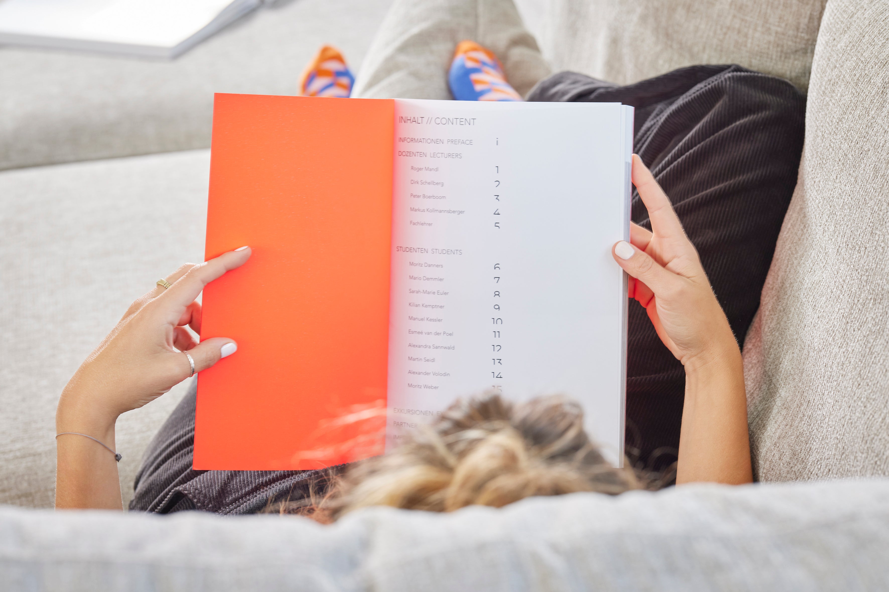 Person liest ein leuchtend orangenes Buch auf einem beigen Sofa von Yvi Living, entspannte Atmosphäre mit bunter Socke im Hintergrund.