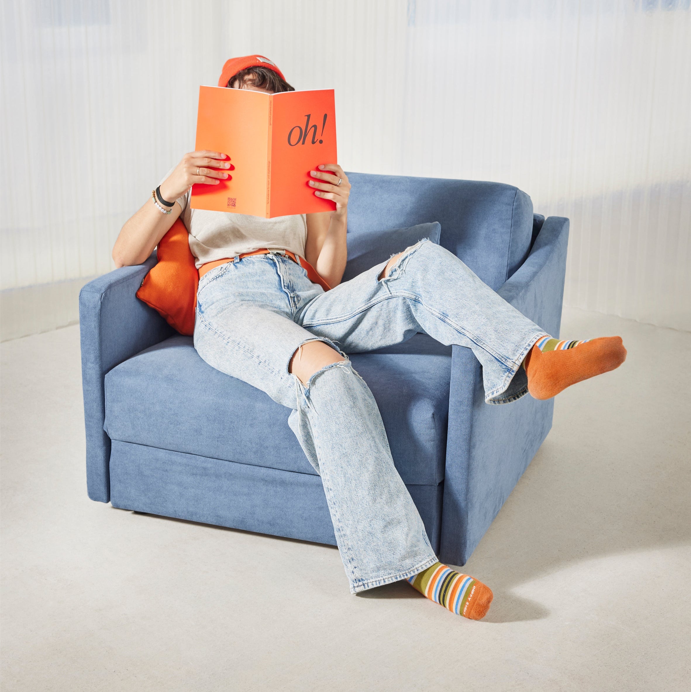 Schlafsessel ERNST in blau steht in einem beigen Raum und wird als Sessel genutzt. Auf dem Sessel sitzt eine Frau und liest ein oranges Magazin mit der Aufschrift "oh!".