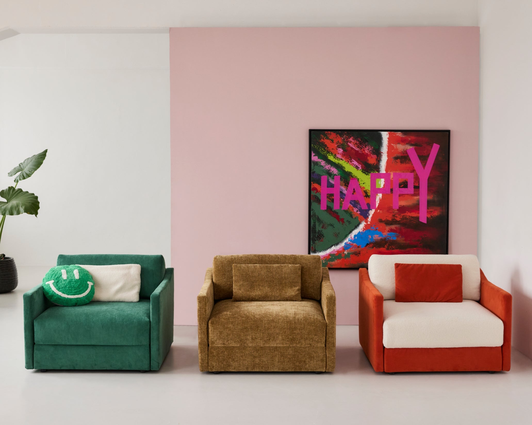 Drei Sessel Ernst von Yvi Living nebeneinander in unterschiedlichen Stoff- und Farbkombinationen:
links Ernst in Teddy Grasgrün, mittig Ernst in Breitcord Goldbraun, rechts Ernst in Teddy Orange mit weißem Teddy-Rücken. Jeder Sessel ist mit passenden Kissen dekoriert. Im Hintergrund eine rosa Wand mit farbigem „HAPPY“-Kunstwerk und links eine große grüne Pflanze. Der Fokus liegt klar auf den drei Stoffvarianten des Sessels Ernst.