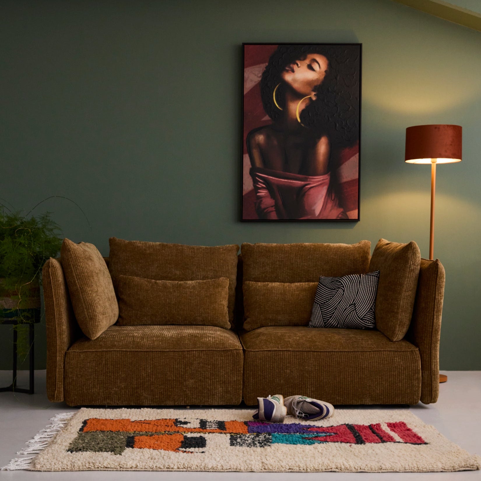 2-Sitzer des Sofas NICA von Yvi Living, bezogen mit Breitcord in Goldbraun, arrangiert vor einer sanft grünen Wand. Das Sofa ist mit passenden Rücken- und Seitenkissen ausgestattet und steht auf einem hellen Boden mit buntem, gemustertem Teppich davor. Eine Stehleuchte und ein Kunstwerk im Hintergrund runden die gemütliche Szenerie ab. Im Mittelpunkt steht das NICA Sofa im Breitcord Goldbraun, das den Raum mit seiner tiefen, weichen Optik prägt.