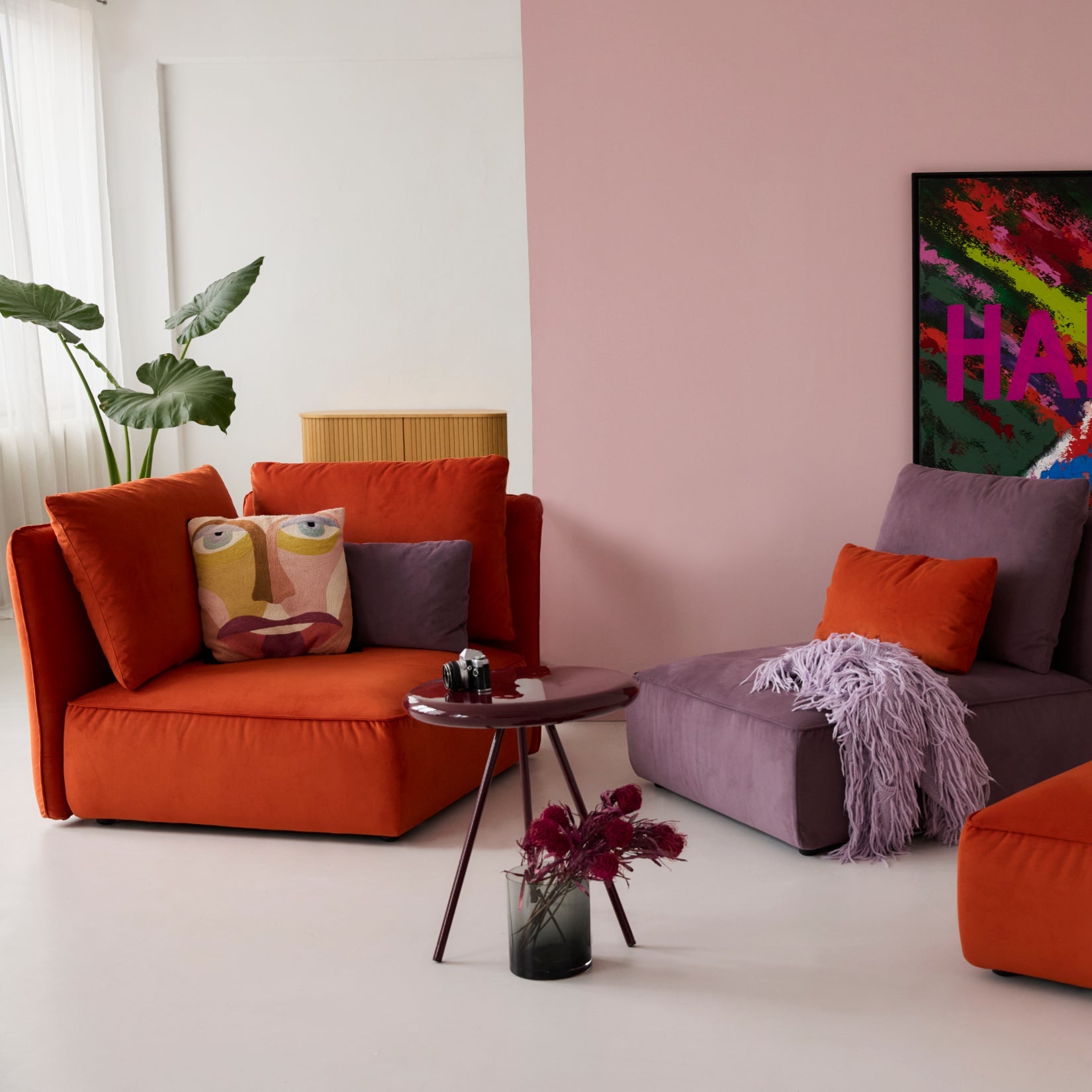 Mehrere Module des Sofas NICA von Yvi Living, bezogen mit Samtstoff in kräftigem Orange und sanftem Violett. Die NICA-Elemente sind als flexible Loungegruppe arrangiert und mit passenden Samtkissen dekoriert. Im Hintergrund ein farbenintensives „HAPPY“-Kunstwerk und links eine große Pflanze. Der Fokus liegt klar auf den NICA Modulen in Samt und ihrer modernen, weichen Optik.